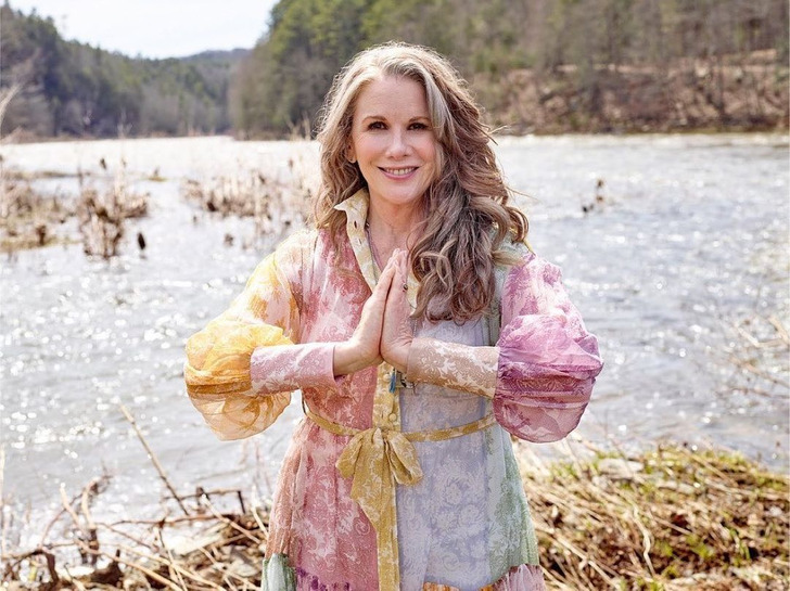 Melissa Gilbert next to a lake wearing colorful clothes and making the namaste sign with her hands.