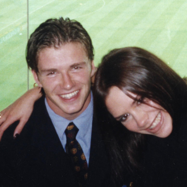 David Beckham in a suit smiles widely as Victoria embraces him, football field in the background.
