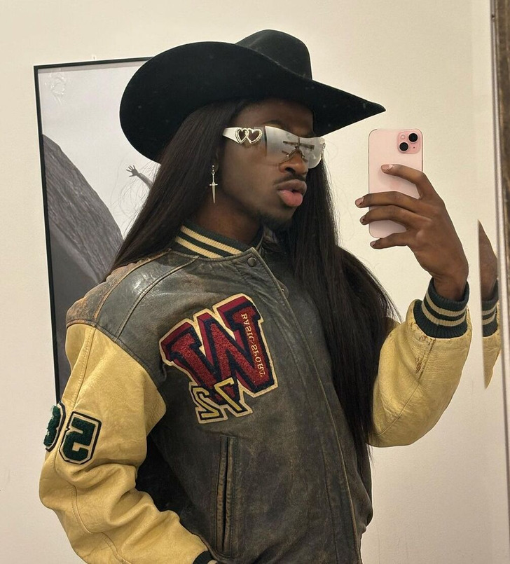 A person with long hair and mustache takes a mirror selfie in a varsity jacket and grey shades.