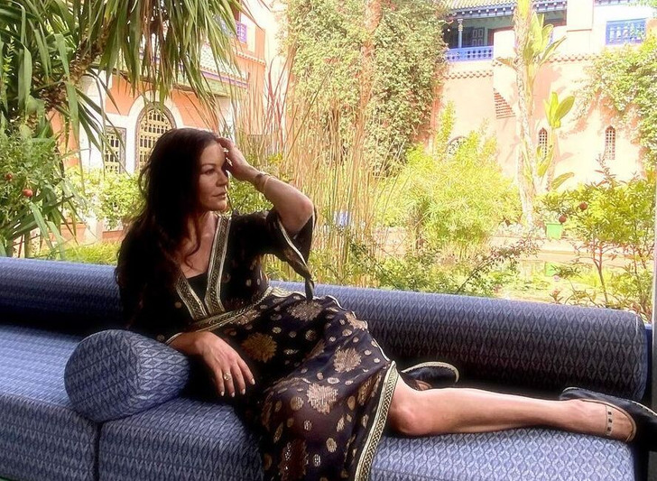 Catherine Zeta-Jones poses on a sofa wearing black tunic, garden in background.