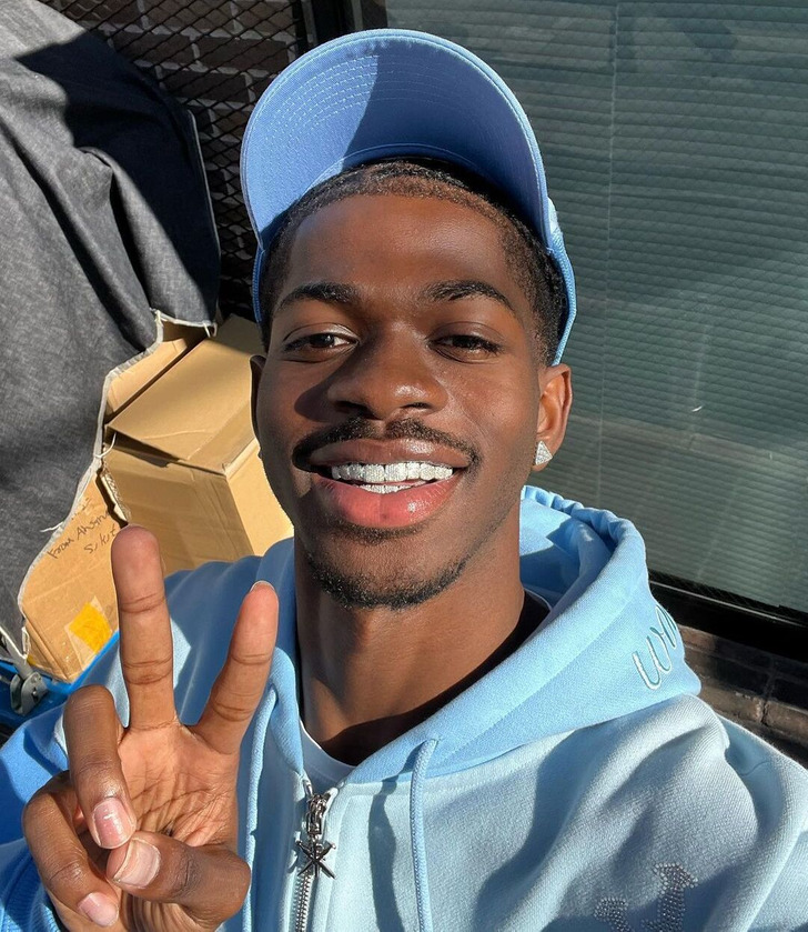 Closeup of Lil Nas X smiling with teeth, making a V sign with his hand, wearing blue hoodie.