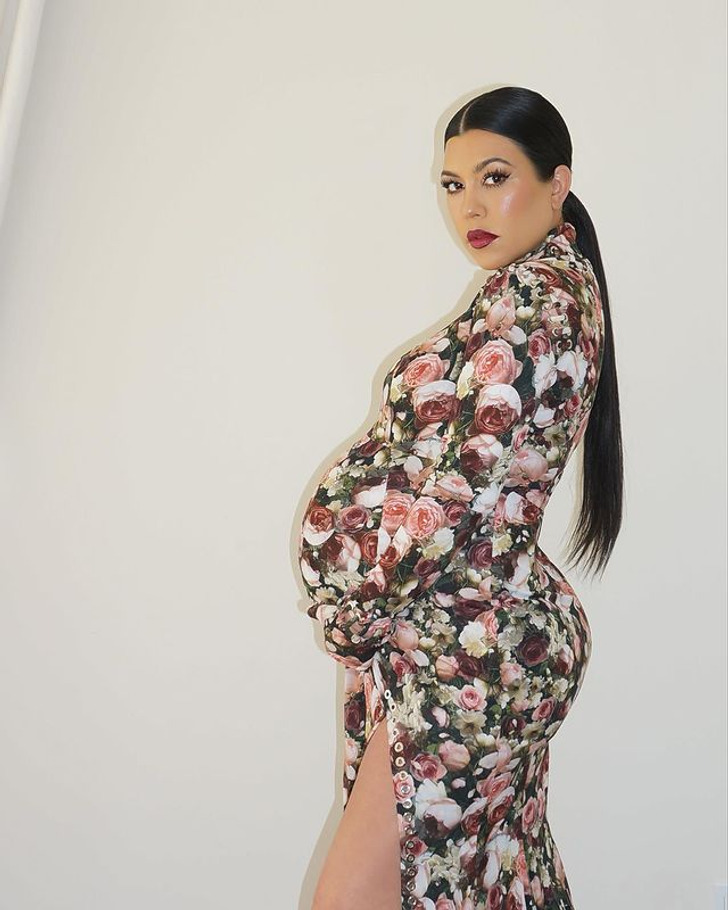Pregnant Kourtney Kardashian wearing a high slit dress with roses, hair in a ponytail.