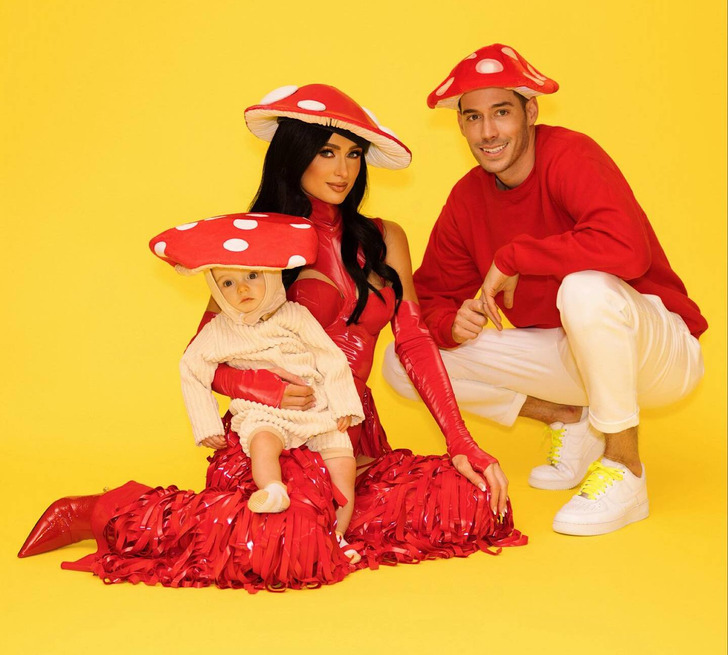 Paris Hilton sitting wearing a red dress holding her son beside her husband, all in mushroom hats.