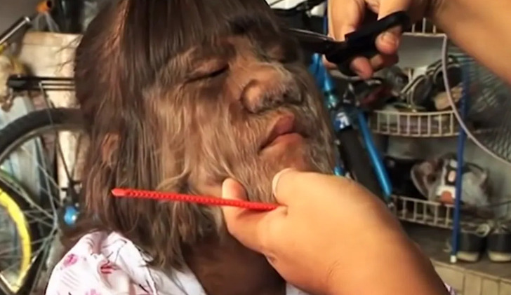 A little girl getting a hair cut, her face covered in fur-like hair, cluttered background.
