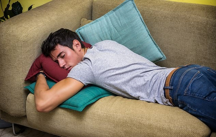 A young man in a grey tshirt and blue jeans sleeping on a couch, resting on cushions.