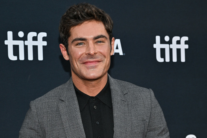 Closeup of actor Zac Efron smiling in a grey suit and black shirt at tiff.