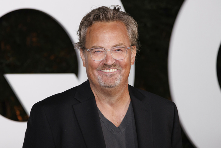 Matthew Perry wearing glasses and a black blazer with grey t shirt.
