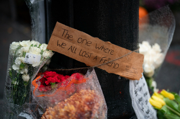 A sign on a post surrounded by flowers that reads "The one where we all lost a friend".