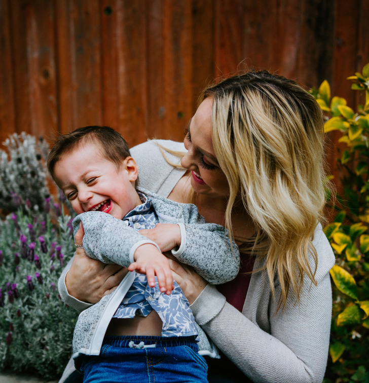 A woman in grey smiles holding a laughing young boy in the backyard.