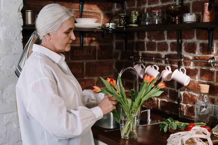 An older woman with grey hair taking care of vase of flowers in her kitchen counter.