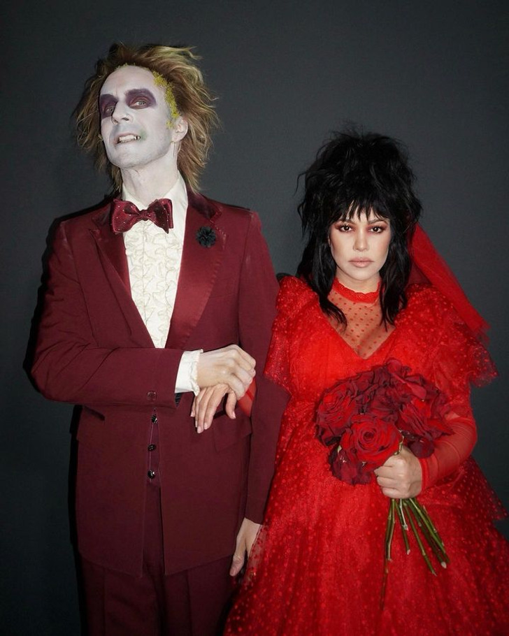 Travis Baker in a maroon suit with face paint and Kourtney Kardashian in a red dress holding roses.
