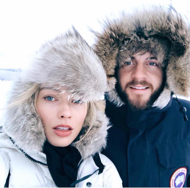 Close up of Margot Robbie and a man wearing winter coats and hats, looking at camera.