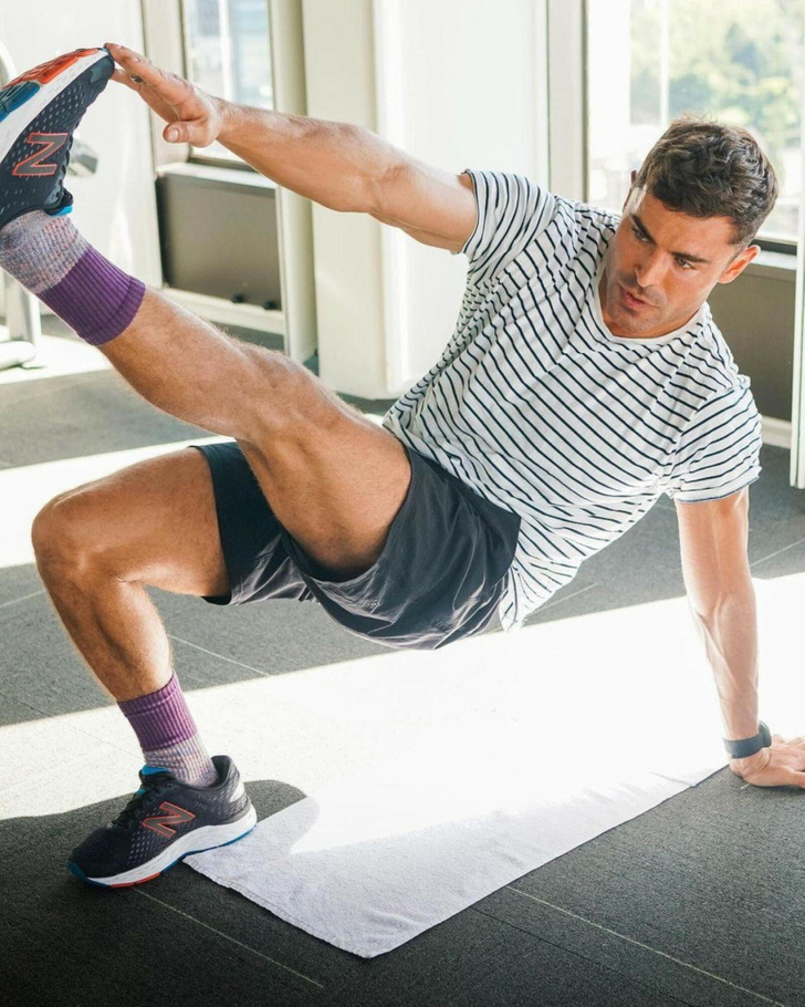 Zac Efron raising his feet and touching it with his hand, wearing white tshirt and black shorts.