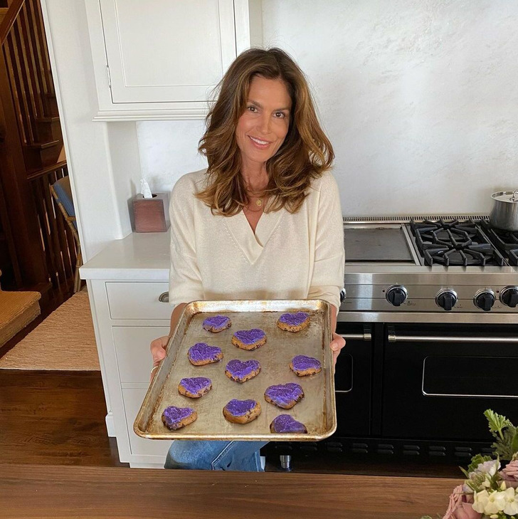 Cindy Crawford holding a baking tray with heart-shaped cookies in kitchen wearing white v neck top.