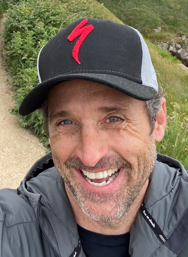 Closeup of Patrick Dempsey smiling widely, wearing a black cap and grey jacket, hilly background.