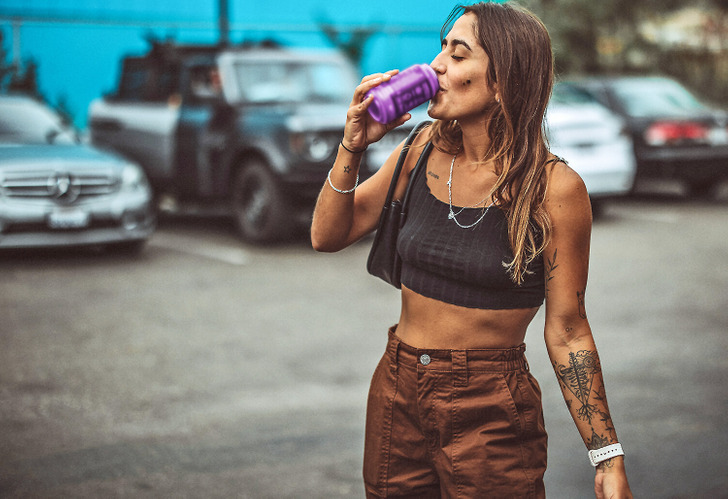 A young tattooed woman in a bra top and cargo pants sipping from a can, cars in the background.