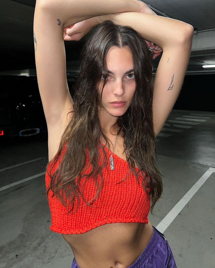 Young brunette woman posing with her arms up in a parking lot and wearing a red crochet tank top.