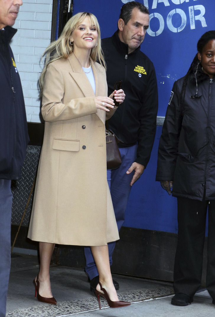 Reese Witherspoon smiles in a long beige coat and brown heels surrounded by security.