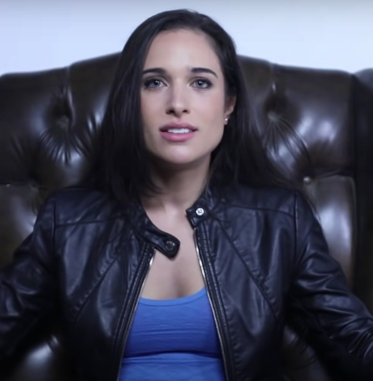 Brunette woman sitting on a black leather chair wearing a black leather jacket and blue shirt.