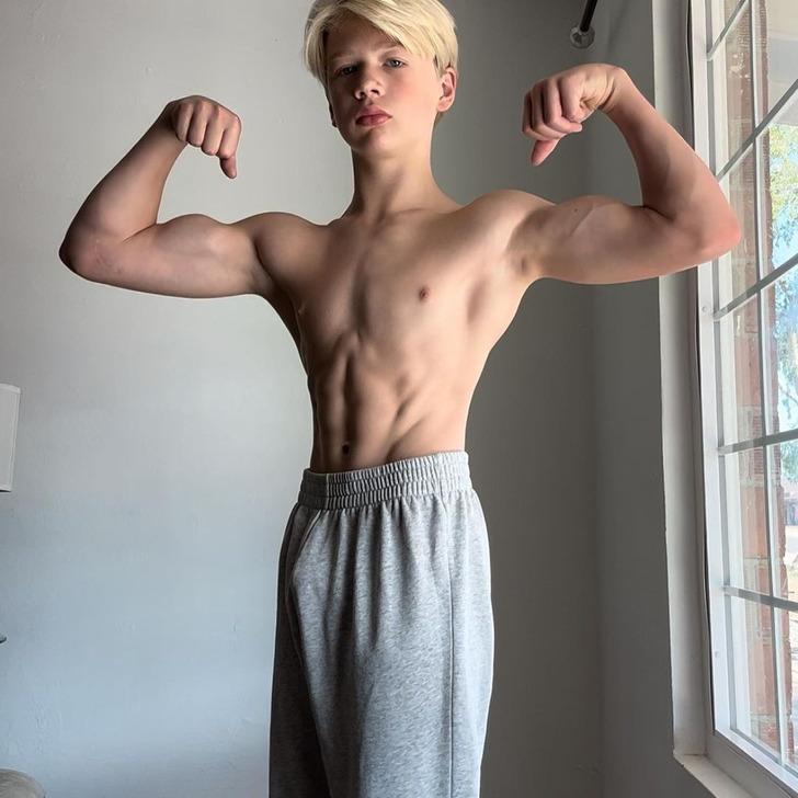 A young boy flexing his arms muscles wearing grey sweatpants.