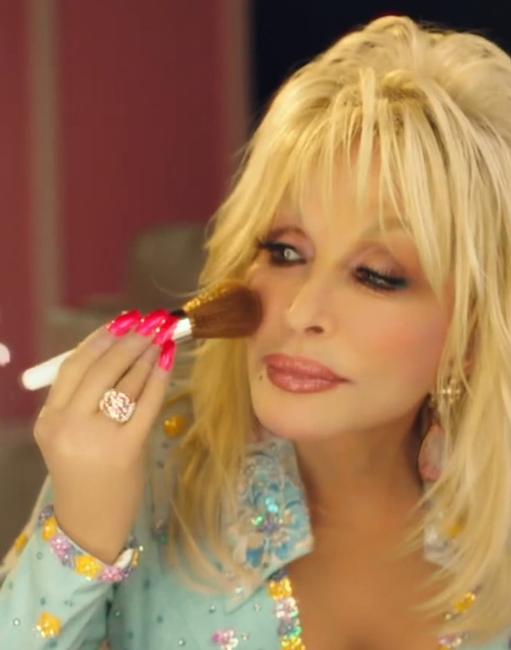 Singer Dolly Parton applying makeup on to her face with a fluffy brush in a blue sparkly outfit.