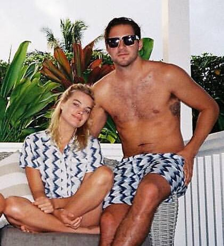 Margot Robbie sits on a sofa with crossed legs, a man beside her with matching clothing.