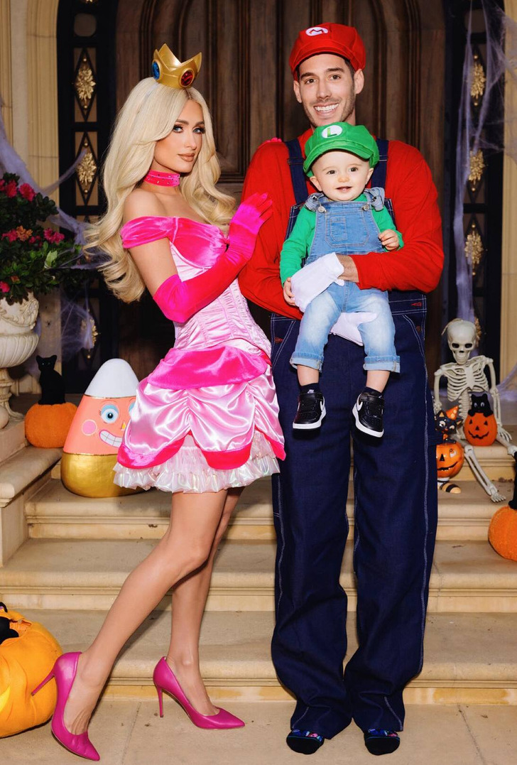 Paris Hilton in hot pink dress, husband in red tshirt and blue dungarees hold son, Halloween decor.