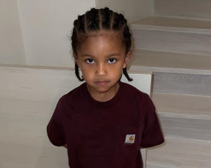 Young boy staring into the camera with his arms behind his back and braided hair.