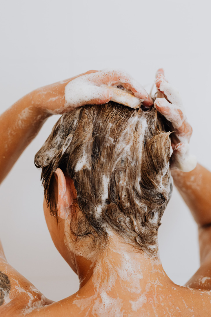 Closeup of a backside of a tattooed woman shampooing her hair, foam around neck and arms.