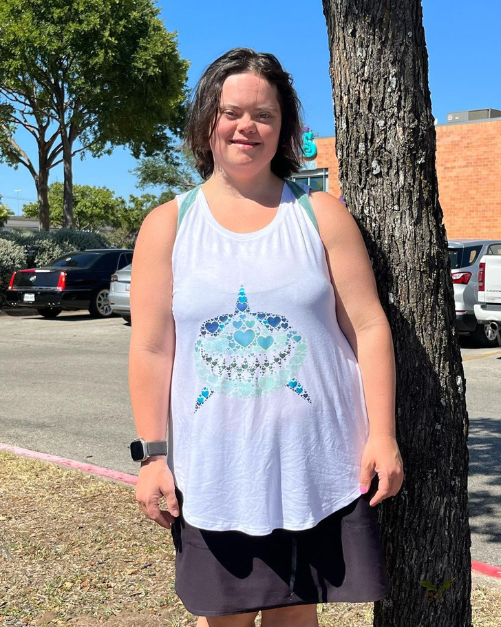 A person with Down Syndrome standing next to a tree in a white tank top and black skirt.