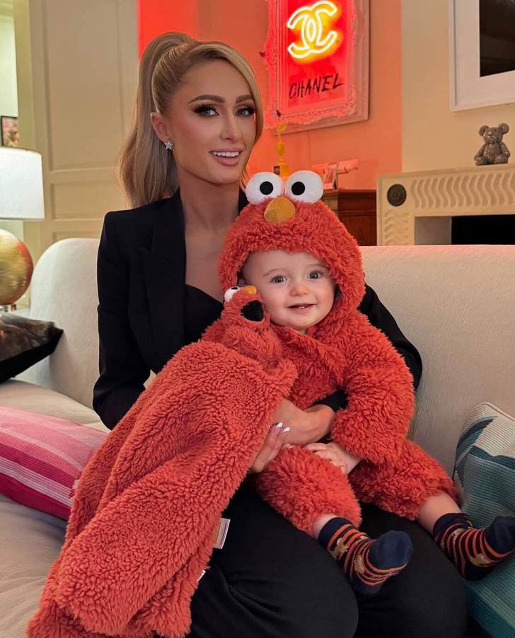 Paris Hilton wearing a black suit, holding her son in Elmo costume, smiles on a couch.