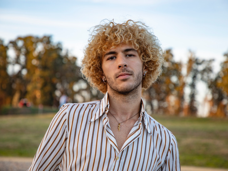 A young man with blonde curly hair looking at the camera, wearing white shirt with stripes.