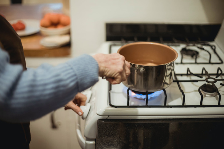 Woman wearing long sleeves blue sweater heating water in a pan on burning stove.