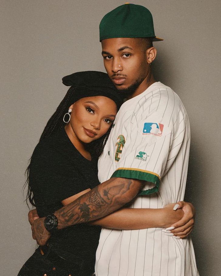 Halle Bailey in black top and headpiece hugs a tattooed man.