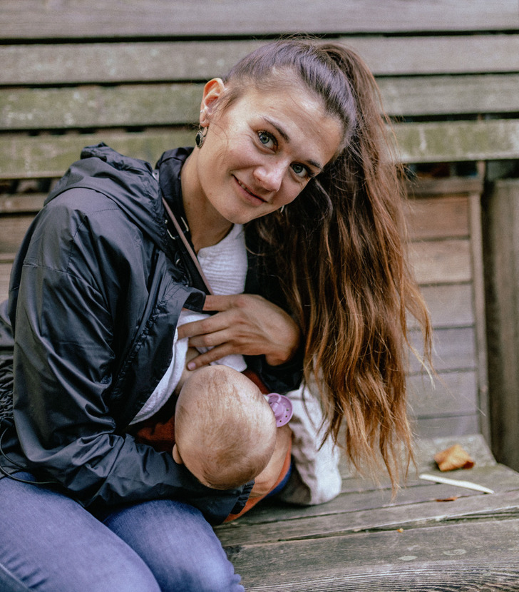 Woman in casual clothes looks at the camera while breastfeeding on a wooden bench.