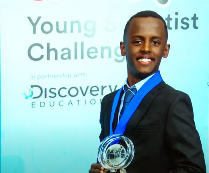 A young boy in a black suit and white shirt wearing a medal and holding a trophy, smiles.