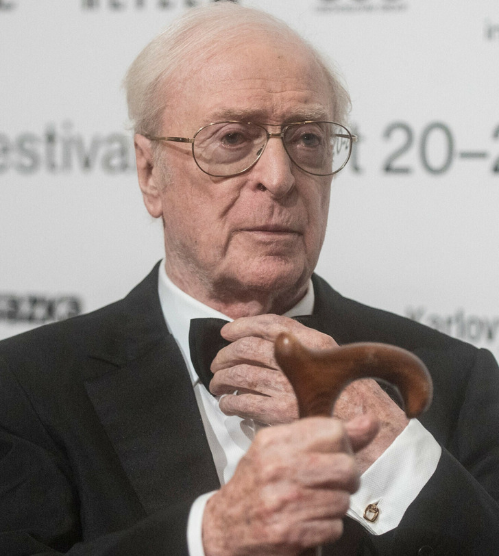 Sir Michael Caine in a black suit, white shirt, holding a cane.