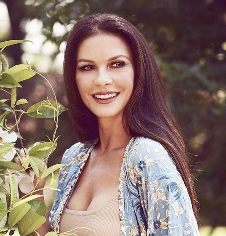 Catherine Zeta-Jones smiling widely in a garden, wearing a beige top and flowery blue shrug.
