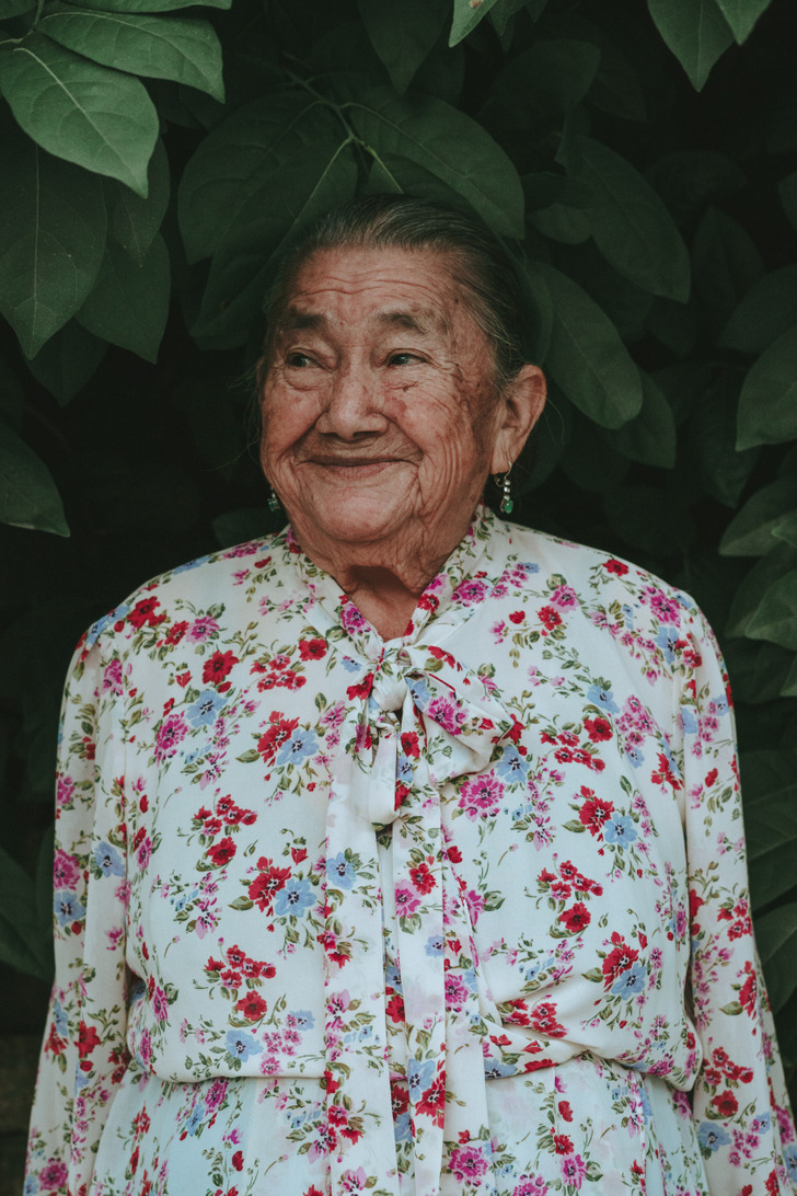 An old woman smiling in front of leaves in a white floral dress.