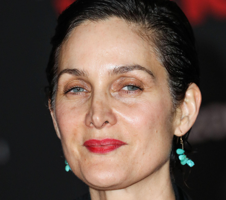 Close-up of Carrie-Anne Moss in black coat, red lipstick, turquoise earring.