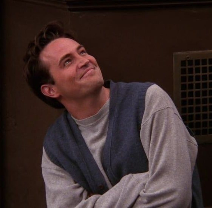 A scene of the tv show Friends with Chandler with arms crossed and wearing grey sweatshirt and vest.