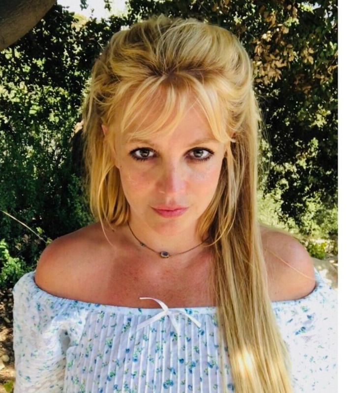 Britney Spears looking at the camera in a white crop off-shoulder top, greenery in the background.