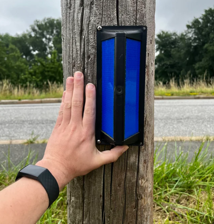 A hand holding a blue light on a wooden pole, roadway in the background.