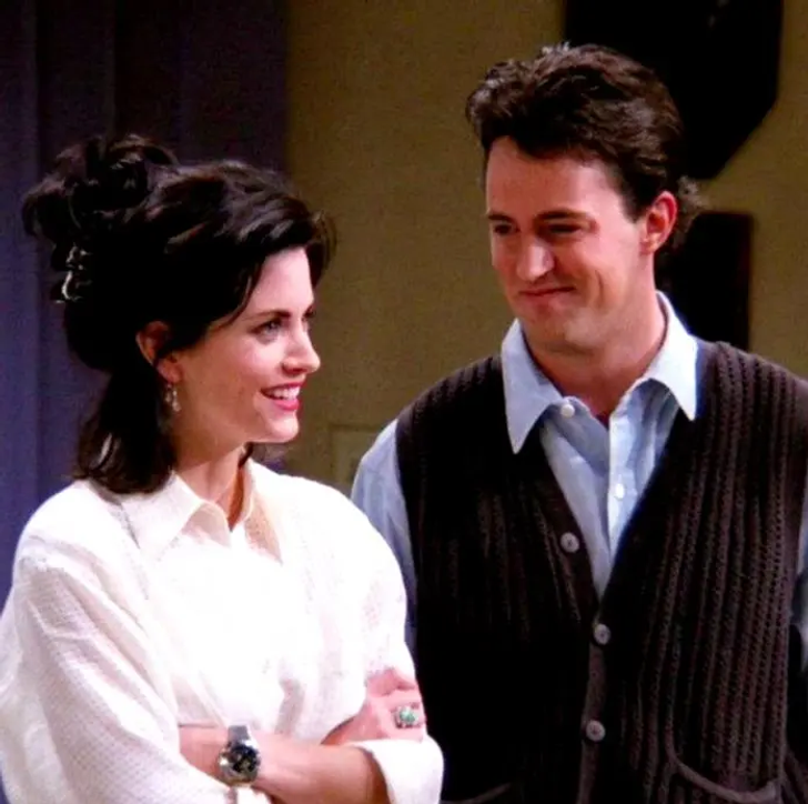 Chandler Bing wearing a grey vest looking at Monica Geller who is wearing a white shirt.