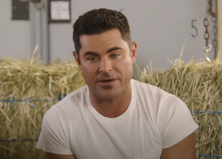 Close up of Zac Efron in a white shirt, stacks of hay in the background.